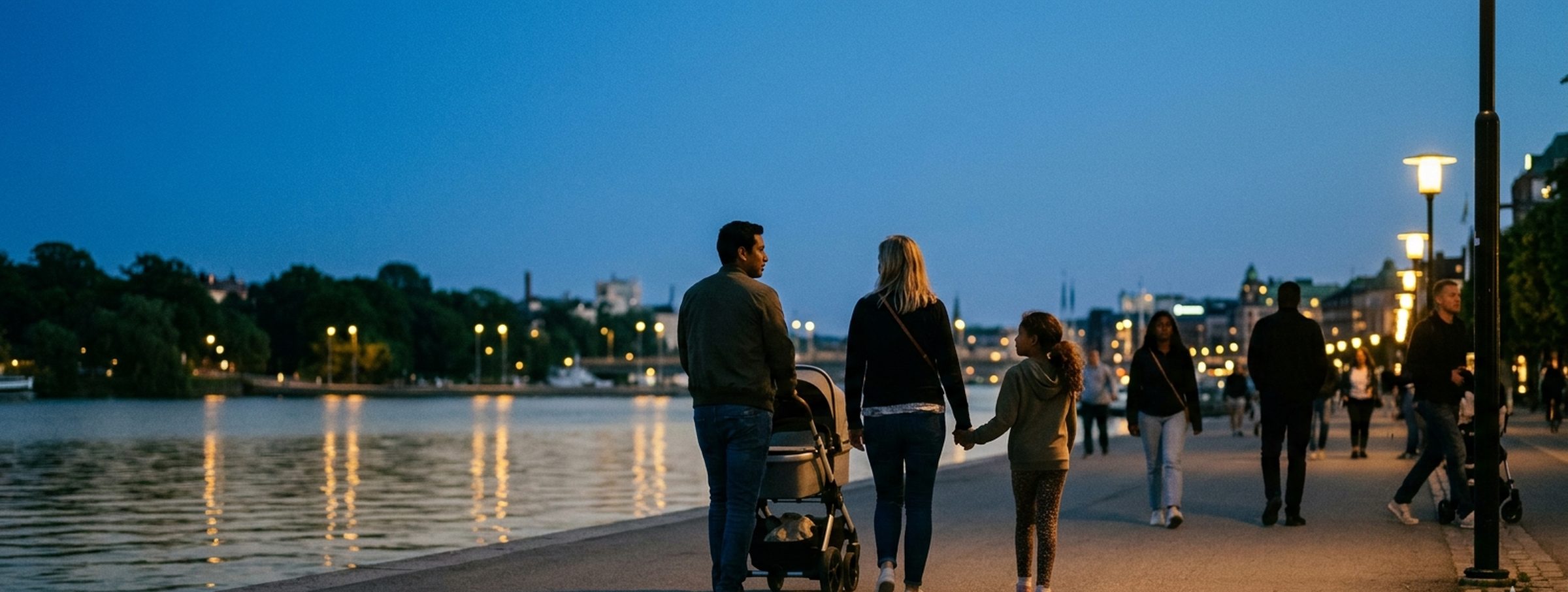 En familj promenerar längs en svensk sjökaj i skymningen, varma stadsljus speglas i vattnet.